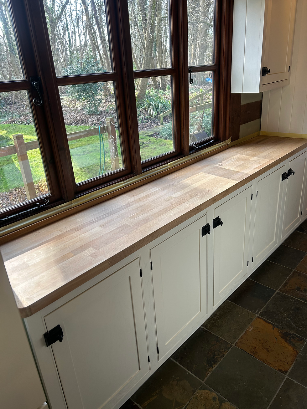 Wooden butcher block countertop above white cabinets facing a large dark wood window overlooking a garden