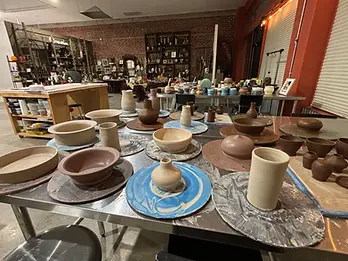 A table is covered with pottery in progress, newly thrown forms still on the bats. The gift shop is in the background.