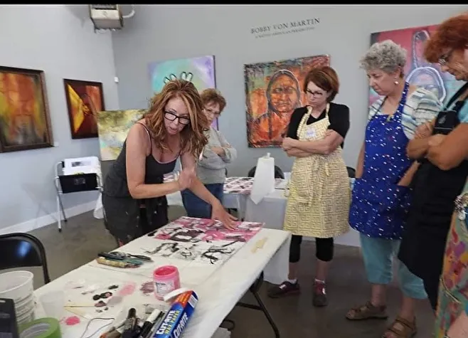 Students are gathered around a table watching Jeannette Goulart teaching a painting class.