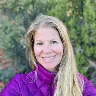Wearing a striking purple colored jacket, Bethany is standing in front of a wall of leaves.