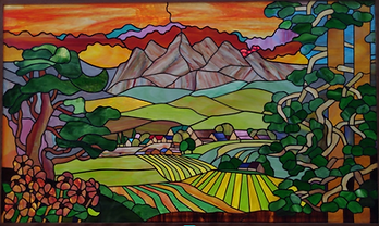 This stained glass window is made of brightly colored glass. The foreground shows flowers and vines on the sides. Criss-crossing fields are in the horizon, with village buildings and a layered mountain range behind.
