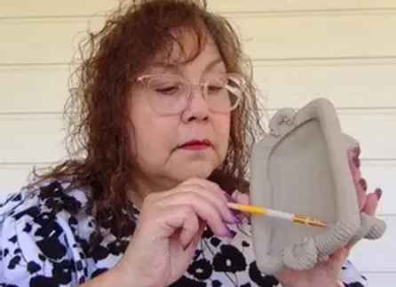 Dinorah Benavides is holding a raw clay platter she's working on with a small pottery tool.