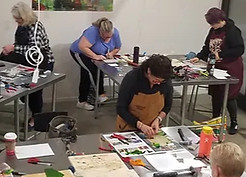 people are shown standing at tables in the classroom, working on art projects.