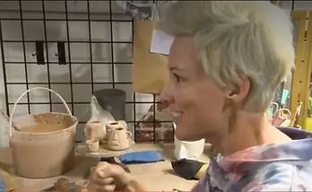 We see a profile view of Raquel by a work bench with clay products and tools on it.