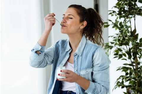 Woman in denim shirt enjoying yogurt with a spoon, eyes closed, smiling. Indoor setting with a green plant in the background.