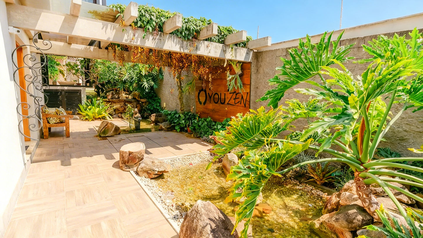 Espaço zen e paisagístico ao ar livre visto sob um ângulo amplo, destacando a transição entre o piso amadeirado, o jardim de pedras e a rica vegetação tropical com destaque para uma grande folhagem de guaimbê em primeiro plano, projeto feito pela RM Arquitetura em Atibaia