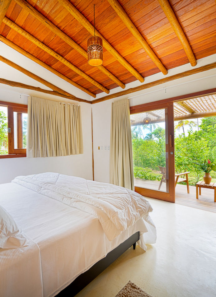 Interior de um quarto de casal iluminado em estilo rústico, com cama confortável e enxoval branco. Destaca-se o teto inclinado com forro e vigas de madeira, além de uma grande porta de correr de vidro que se abre para uma varanda privativa com deck, cercada por um jardim tropical com coqueiros ao fundo. Nas casas da Vida Cajuína, localizada na Praia do Cassange, em Maraú, Bahia.
