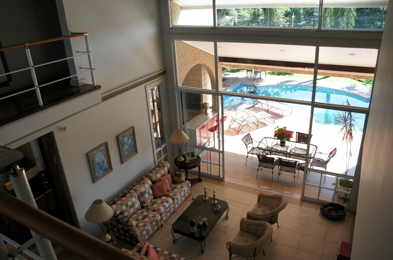 Vista superior de ampla sala de estar com pé-direito duplo, mezanino e grandes painéis de vidro que integram perfeitamente o ambiente interno à varanda coberta e à área da piscina feito pela RM Arquitetura