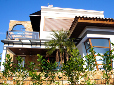 Fachada contemporânea de residência de alto padrão com dois pavimentos, apresentando revestimentos em pedra natural, ampla varanda superior com guarda-corpo de vidro, esquadrias em tom amadeirado e paisagismo frontal com palmeira e folhagens tropicais.