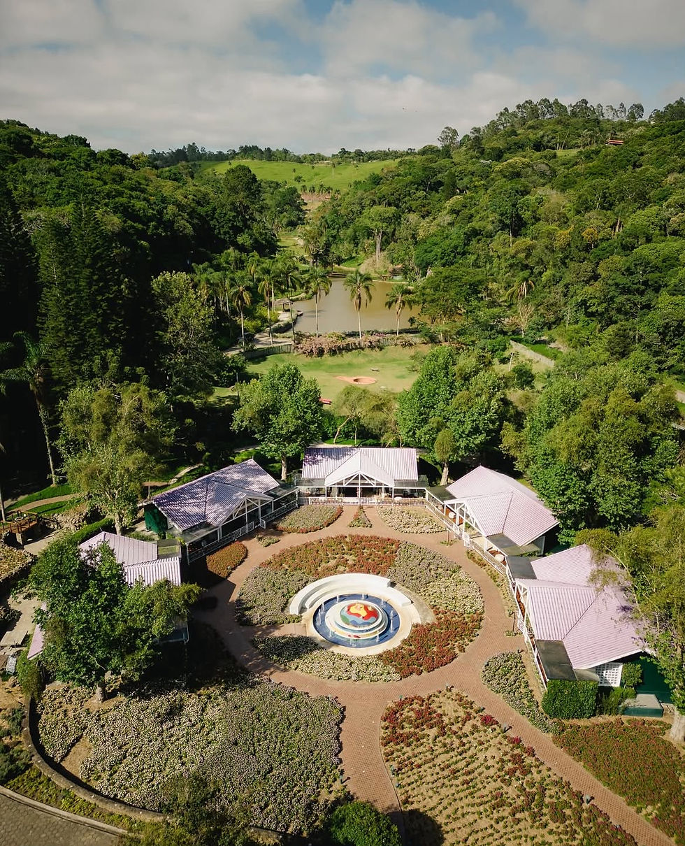 Vista aérea espetacular de uma vila de chalés dispostos de forma semicircular em volta de uma praça central bem desenhada. No centro da composição sobressai uma fonte ornamental em formato circular, cercada por canteiros de flores geométricos e caminhos bem definidos