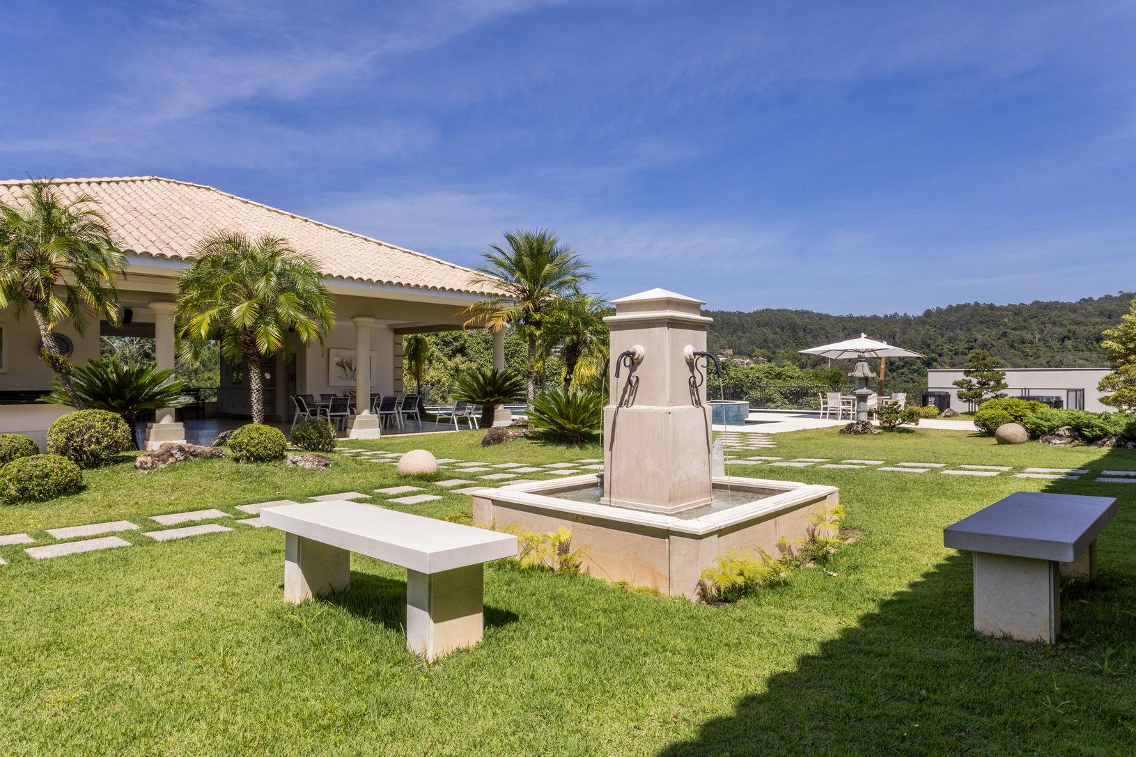 Jardim clássico com fonte de água central, bancos de pedra, gramado impecável e palmeiras em torno da residência.Excelente exemplo de Arquitetura em Atibaia focada no paisagismo elegante e na valorização da área externa feito pela RM Arquitetura.