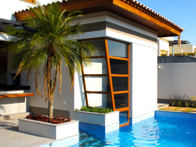 Área externa contemporânea com piscina de água azul, integrada a anexo com grande painel de vidro e caixilharia de madeira inclinada, palmeira em canteiro elevado e acabamentos de alto padrão.Projeto moderno assinado por Arquiteto em Atibaia, combinando design arrojado, elementos naturais e integração com o espaço de lazer.