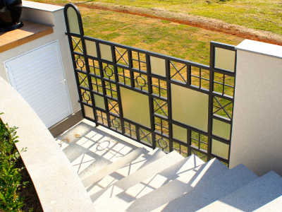 Portão de acesso externo e guarda-corpo com design geométrico exclusivo em serralheria preta e painéis de vidro opaco, instalado ao lado de escada externa de concreto.