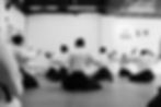 Foto panorâmica em preto e branco de alunos de costas praticando um exercício de abertura de braços e respiração sentados no aikido adulto em pinheiros nas aulas do En Dojo. 