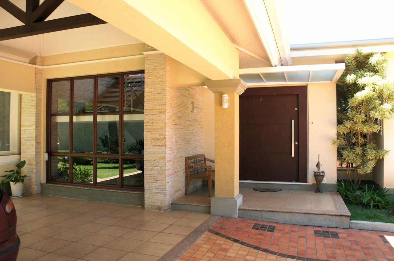 Entrada principal de residência elegante com imponente porta pivotante em tom escuro, puxador vertical metálico, alpendre com coluna em tom amarelo e parede revestida em pedra canjiquinha feito pela RM Arquitetura em Atibaia