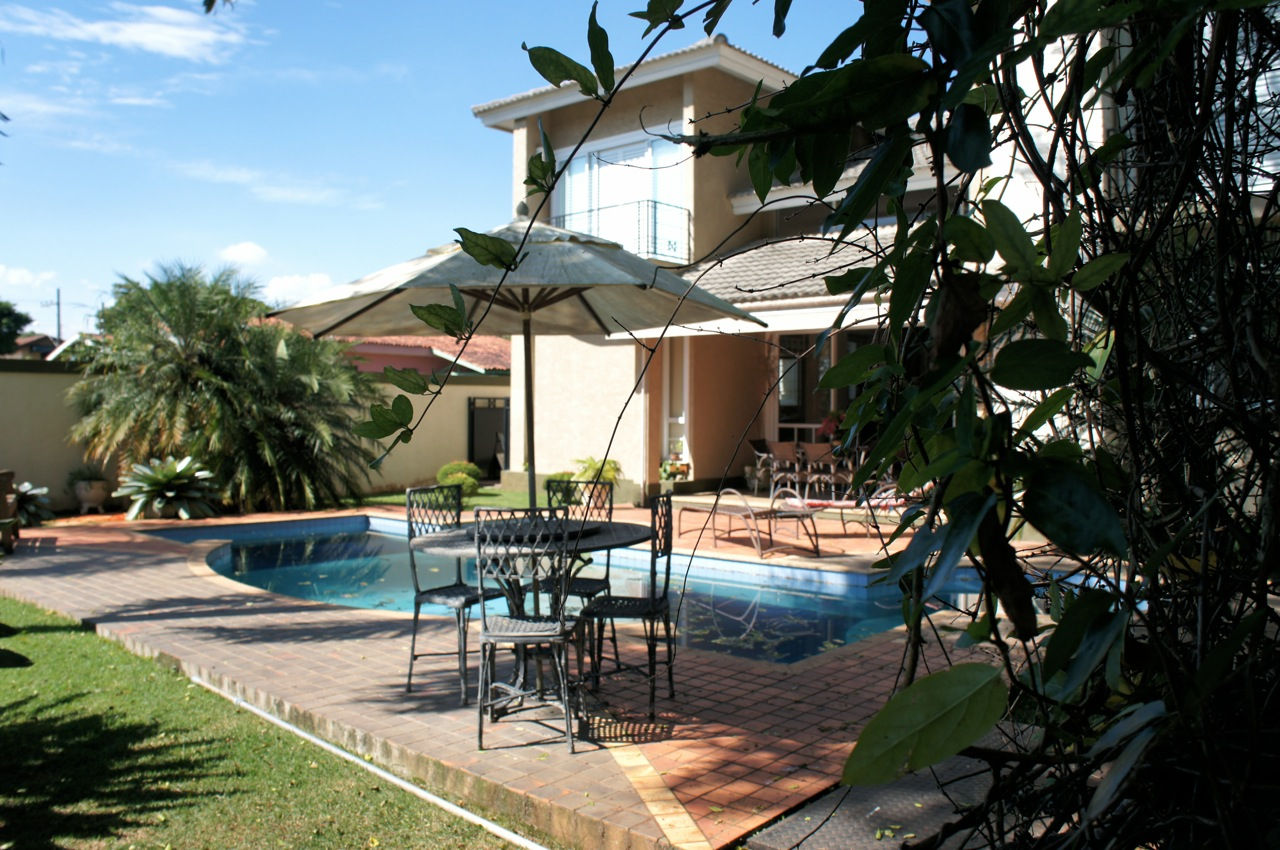 Área externa de lazer com piscina de desenho misto, deck cerâmico, mesa de apoio com ombrelone, jardim com vegetação tropical e fachada posterior de residência de dois pavimentos