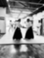 Foto em preto e branco de um treino de Aikido no dojo de Aikido En Dojo, em Pinheiros. No primeiro plano, uma praticante e um praticante de hakama preto seguram o punho um do outro, com outros alunos treinando no fundo.