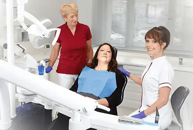 Das Bild zeigt eine Zahnklinik mit drei Frauen. Eine Frau, die ein blaues Lätzchen trägt, sitzt auf einem Zahnarztstuhl und scheint eine Patientin zu sein. Eine andere Frau in einem burgunderroten Kittel steht neben ihr, während eine dritte Frau in einem weißen Kittel mit lila Handschuhen neben der Patientin sitzt und in die Kamera lächelt. Die Szene zeigt typische zahnärztliche Geräte wie eine Deckenlampe, Tabletts und zahnärztliche Instrumente. Es sind keine erkennbaren Figuren, Orientierungspunkte oder Marken zu sehen.