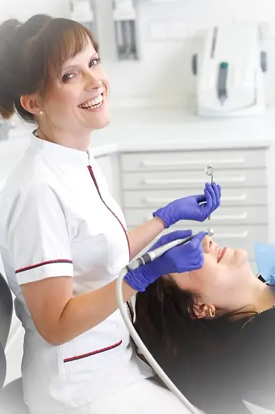 Das Bild zeigt eine freundliche Zahnärztin oder Dentalhygienikerin in weißer Uniform mit roter Verzierung, die in die Kamera lächelt. Sie trägt lila Handschuhe und hält zahnärztliche Instrumente in der Hand. Neben ihr lehnt sich eine Frau in einem Zahnarztstuhl zurück und bereitet sich auf einen zahnärztlichen Eingriff vor, wobei sie ein zahnärztliches Lätzchen trägt. Der Hintergrund besteht aus der typischen Einrichtung einer Zahnarztpraxis, einschließlich weißer Schränke, Schubladen und zahnmedizinischer Geräte. Auf dem Bild sind keine erkennbaren Figuren, Orientierungspunkte oder Marken zu sehen.