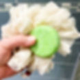 Hand holding a natural shower puff made of ramie and a green shampoo bar with a shower enclosure in the background