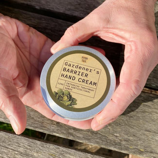 Two hands holding a tin of Gardener's Barrier Hand Cream over a garden table
