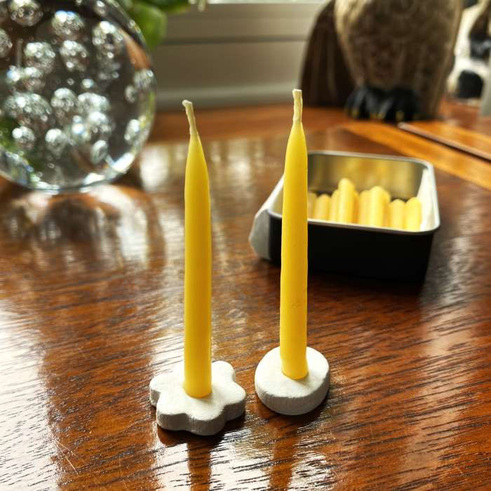 Two handmade mini taper candles standing in handmade clay holders with their storage tin in the background