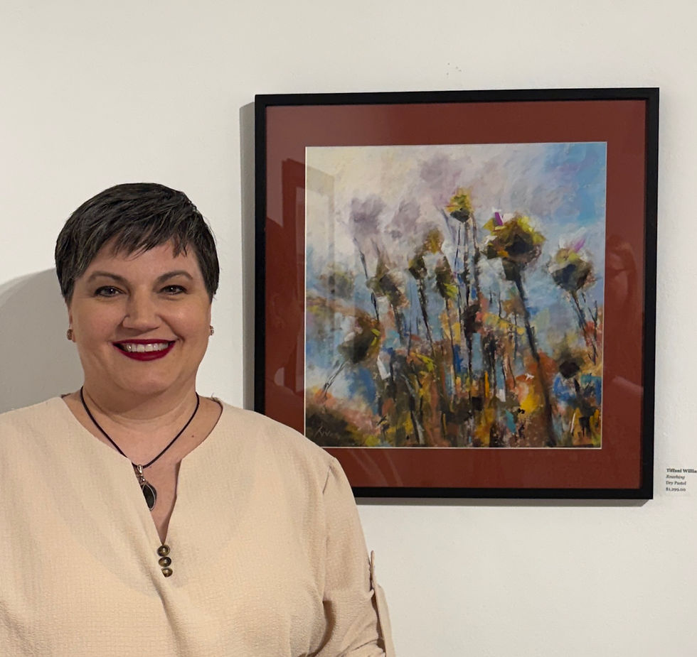 Tiffani Williams smiles as she poses in front of her framed artwork "Reaching" which depicts a closeup of weeds and white flowers reaching towards a sunny blue sky.