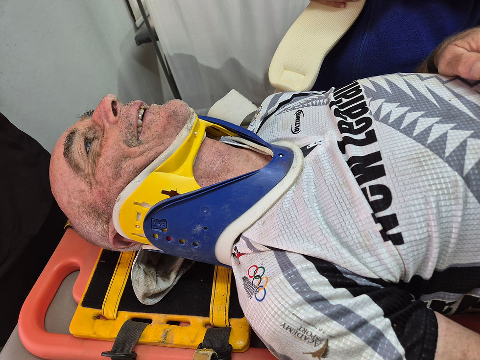 Injured cyclist lies on a stretcher wearing a blue-and-yellow neck brace and white team jersey, looking up and smiling.