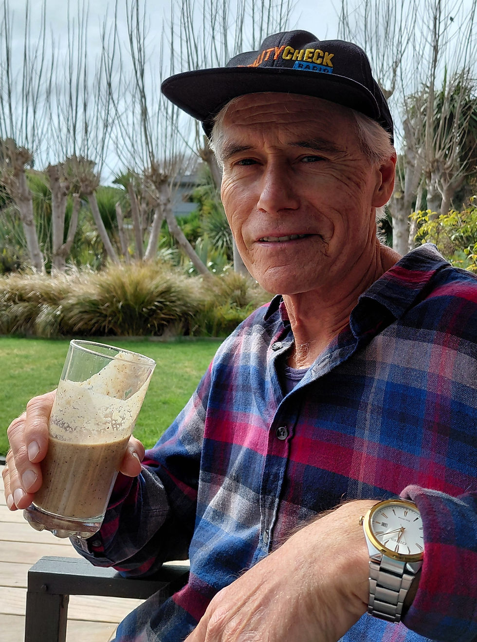 Elderly man in a plaid shirt and "BeautyCheck" hat holds a smoothie. He sits outdoors with leafless trees in the background, looking relaxed.