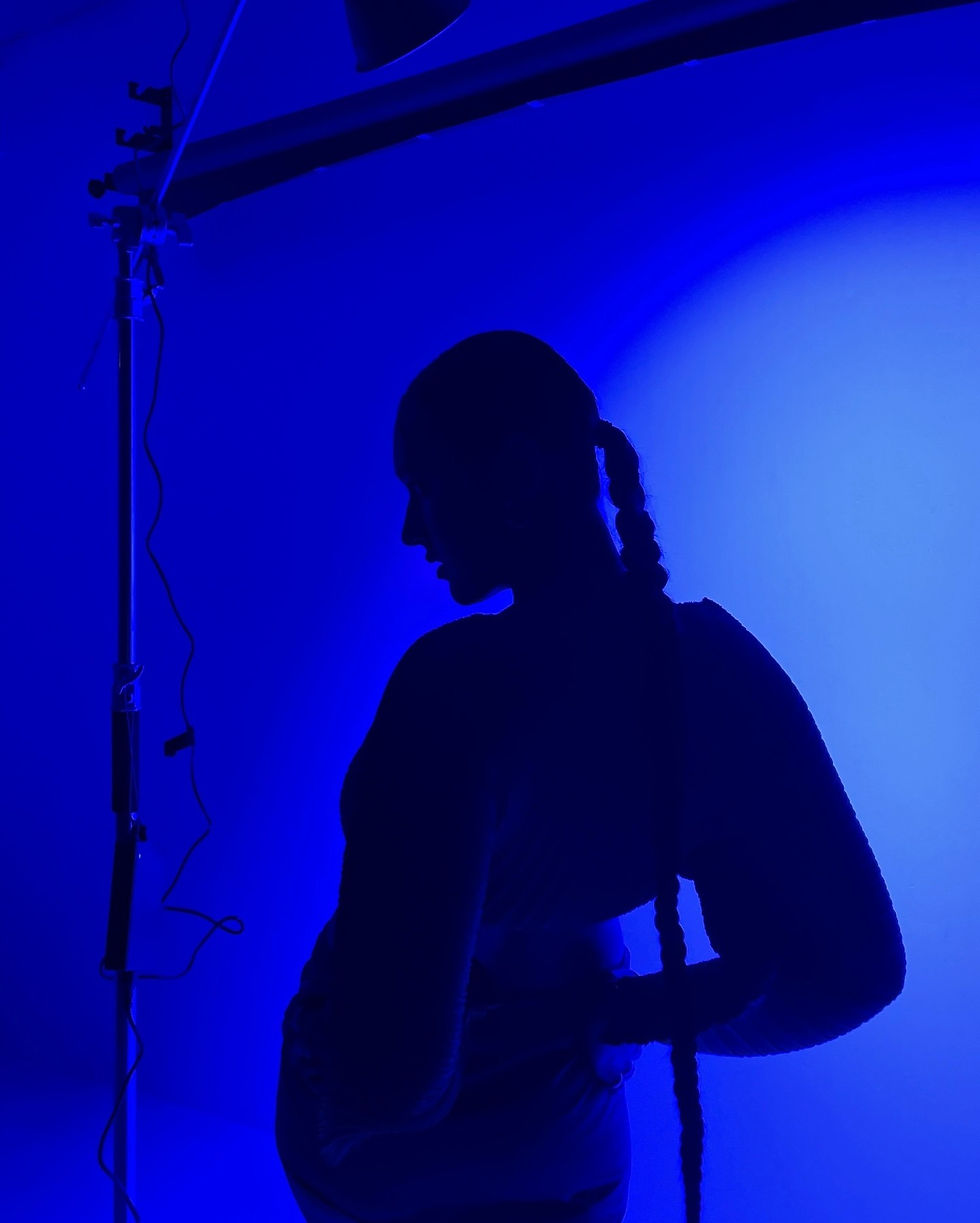 Silhouette of a person with braided hair in blue lighting, standing with hand on hip. Studio setup visible, creating a mysterious mood.