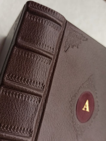 Close-up of a custom bound book in brown goat leather with raised bands on the spine, detailed blind tooling, and a red leather onlay with a gold monogram “A”.