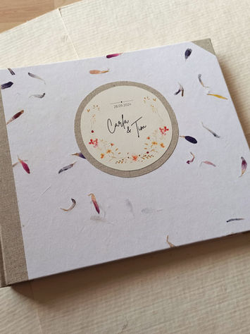 Handcrafted custom wedding book bound in natural bookbinding cloth, featuring handmade paper with dried flower petals and a personalized title plate created from the couple’s wedding invitation.