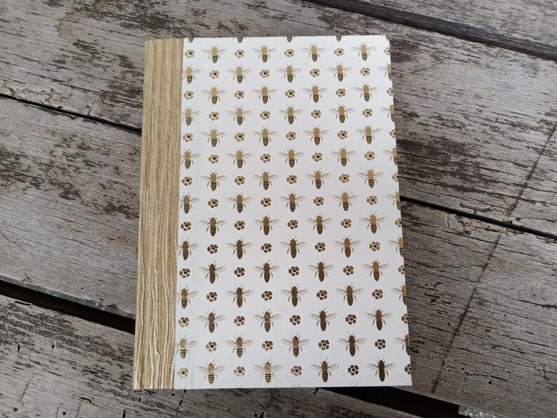 Handgebundenes Imkerjournal mit weißem Einband in goldenen Bienenmotiven und strukturiertem goldenem Rücken.