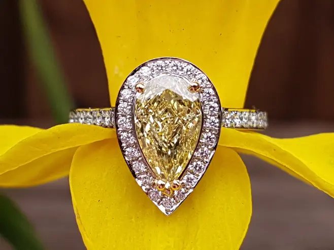 The ring shows a platinum and diamond set ring, with a pear-shaped yellow diamond at the centre. The ring has been photographed sitting on a big, bright yellow daffodil.