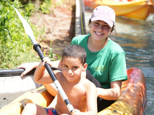 A young boy and a woman in a green shirt and pink cap are kayaking on a calm body of water. The boy, shirtless and wearing red shorts, holds a paddle with both hands, while the woman sits behind him, smiling. They are in a yellow kayak, with other kayaks and greenery visible in the background under a bright, sunny sky.