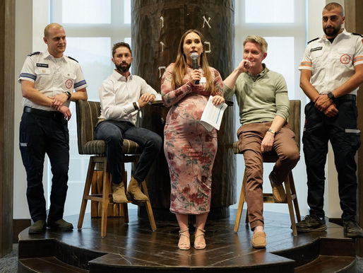 A group of five people is on a small stage with a wooden backdrop featuring Hebrew text. A pregnant woman in a floral dress stands in the center holding a microphone and a piece of paper. Two men in casual attire sit on stools on either side of her. Two individuals in paramedic uniforms stand on the outer edges, one on each side. The setting appears to be an indoor event or discussion panel.