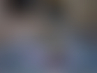 A elegantly set dining table with a teal tablecloth, featuring multiple wine glasses, silverware, and folded napkins. At the center is a gold candelabra with three blue candles emitting a warm glow, surrounded by a floral arrangement of white and purple flowers. In the background, additional tables with similar setups and lit candles are visible, with bookshelves lining the walls of a softly lit room.