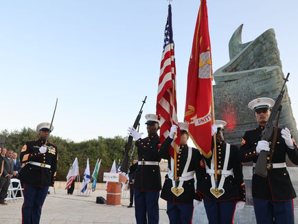 Military personnel in dress uniforms carrying the American flag and a Marine Corps flag, marching in a ceremonial formation. A large abstract monument is in the background, with other flags and people seated or standing in attendance. The scene takes place outdoors under a clear sky.
