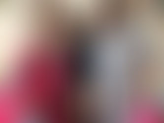 Three smiling women stand together at a community event. The woman on the left wears a red dress and patterned headscarf, holding a pink gift bag with blue tissue paper. The woman in the center wears a black top and patterned skirt. The woman on the right wears a blue and white floral dress and holds a pink gift bag, water cup, and glasses. Behind them are colorful framed artworks and a table with event materials.