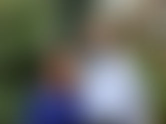 A smiling Orthodox Jewish couple stands outdoors in front of lush greenery and tropical plants. The woman has light brown hair and is wearing a royal blue dress, while the man has a long salt-and-pepper beard and is wearing a light blue button-down shirt. They are standing close together, radiating warmth and joy. The background includes palm leaves, olive branches, and decorative wrought-iron elements.