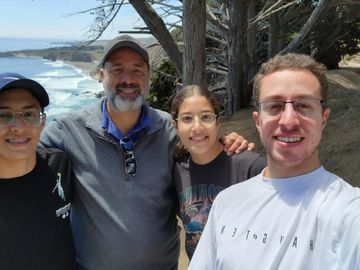 A smiling group of four people, three young adults and one older man—pose for a selfie with their arms around each other in front of a scenic coastal cliff. Behind them are windswept trees and the ocean, with waves crashing along a rugged shoreline. It's a sunny day, and everyone is wearing glasses and casual clothes.