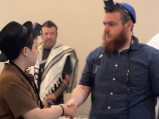 A scene inside a room where two individuals, dressed in traditional Jewish attire including a black hat and a blue kippah, are shaking hands. In the background, another person wearing a prayer shawl (tallit) stands observing. The table in the foreground holds religious items, a book, and a cup, suggesting a communal or religious gathering, possibly related to a prayer or study session.
