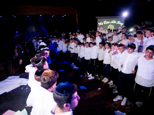 A large group of teenage boys, dressed in white shirts and dark pants with tzitzit, stand arm-in-arm in a circle on a stage during a nighttime event. Many are wearing kippahs. The atmosphere is energetic and joyful, with purple stage lighting and a sign in the background that reads “We Can All Sing.” Girls and other attendees are visible in the dimly lit audience, creating a celebratory and unified scene. Confetti and papers are scattered on the floor, adding to the festive ambiance.