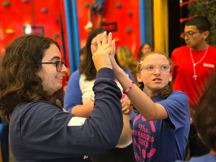 A lively indoor scene at a climbing gym where two young individuals, wearing matching blue Friendship Circle Los Angeles t-shirts, assist each other on a red climbing wall. One person, with glasses and curly hair, reaches up to help the other, who is also wearing a harness and smiling. In the background, other participants and a volunteer in a red shirt observe, with colorful climbing holds and equipment visible, creating a joyful and supportive atmosphere.
