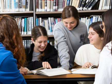 A group of high school girls sit around a table in a library, smiling and studying Jewish texts together. Bookshelves filled with seforim (religious books) line the background, and the atmosphere is warm and collaborative.
