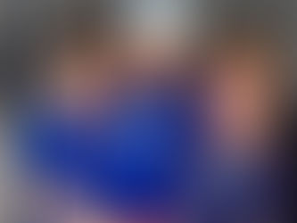 Three young boys stand together smiling in front of a dark metal wall, wearing matching blue "emek early childhood camp" t-shirts. The boy in the center wears a white cap with the same logo, while the others have a palm tree and sun design on their shirts. They are dressed in colorful shorts—light blue, red and white striped, and watermelon-patterned—hugging each other in a joyful group pose.