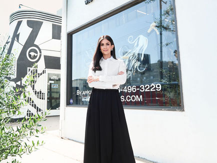 A woman stands with her arms crossed in front of a building with a sign reading "Wig Shop & Salon" and a phone number "4-496-3229" on the window. She is wearing a white blouse, a long black skirt, and black-and-white shoes. The building has a modern design with large windows and a mural featuring abstract black and white patterns on the left side. There is a small tree