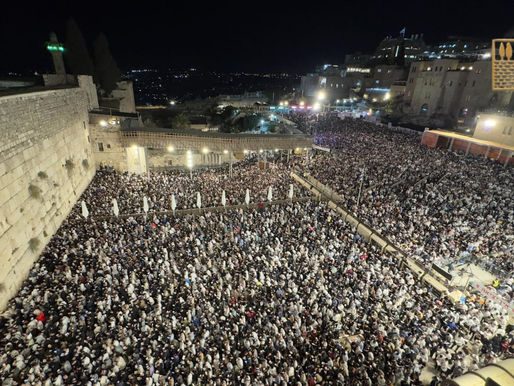 A nighttime aerial view of thousands of people gathered at the Western Wall in Jerusalem for Selichot prayers, with the ancient stone wall on the left and a large crowd filling the plaza. The scene is illuminated by artificial lights, with buildings and a minaret visible in the background, and two emblematic shields featuring Jewish symbols in the top right corner.