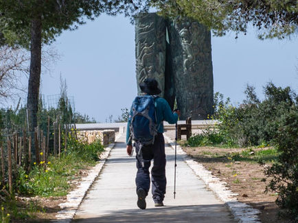 A hiker with a backpack and trekking pole walks along a smooth, paved accessible trail toward the towering bronze Scroll of Fire monument in the Martyrs’ Forest. The path is framed by green pine branches overhead and wildflowers along the edges under a clear blue sky, symbolizing inclusion and remembrance in Israel’s Judean Hills.