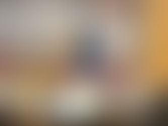 Children’s Hanukkah art display in a classroom: a large painted poster on a dark gray background shows a woman in a blue dress and black bonnet walking through snowy landscape with a basket of oranges, holding a lit torch. In the foreground, a golden branched menorah with glowing orange flames stands prominently. Behind her, a bare tree and full moon complete the winter scene. Below the poster, a table is covered with colorful handmade menorahs, glass bead creations, and other kids’ crafts by students (labeled Raphael 3b, Rina 3b, etc.), with young children in kippot visible in the foreground.